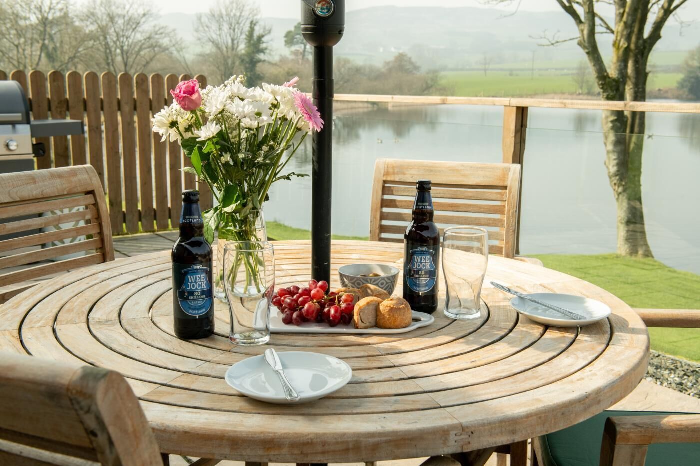 A round wooden table with a bottle of beer, a glass, a plate with bread, and a vase with flowers.