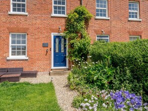 A red brick building with a blue door, a small garden with various plants and flowers, and a gravel pathway. - 2 Bed in Norwich (oc-78491) (Bacton)