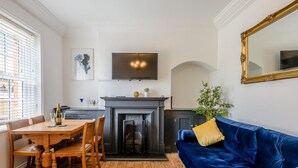 Een woonkamer met een open haard, een blauwe bank, een eettafel met stoelen, en een televisie boven de haard.