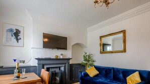 Een woonkamer met een open haard, een blauwe bank, een eettafel met stoelen, en een televisie boven de haard.