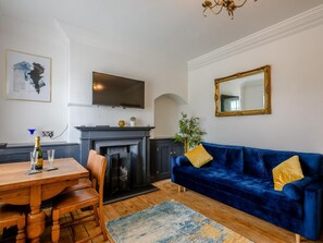 A living room with a fireplace, a blue sofa, a dining table with chairs, and a television mounted above the fireplace. - 2 Bed in Norwich (oc-78491) (Bacton)
