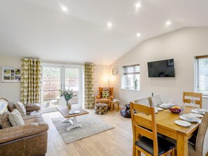 A spacious living area with a sofa, a dining table set for four, a television mounted on the wall, and a large window with curtains.