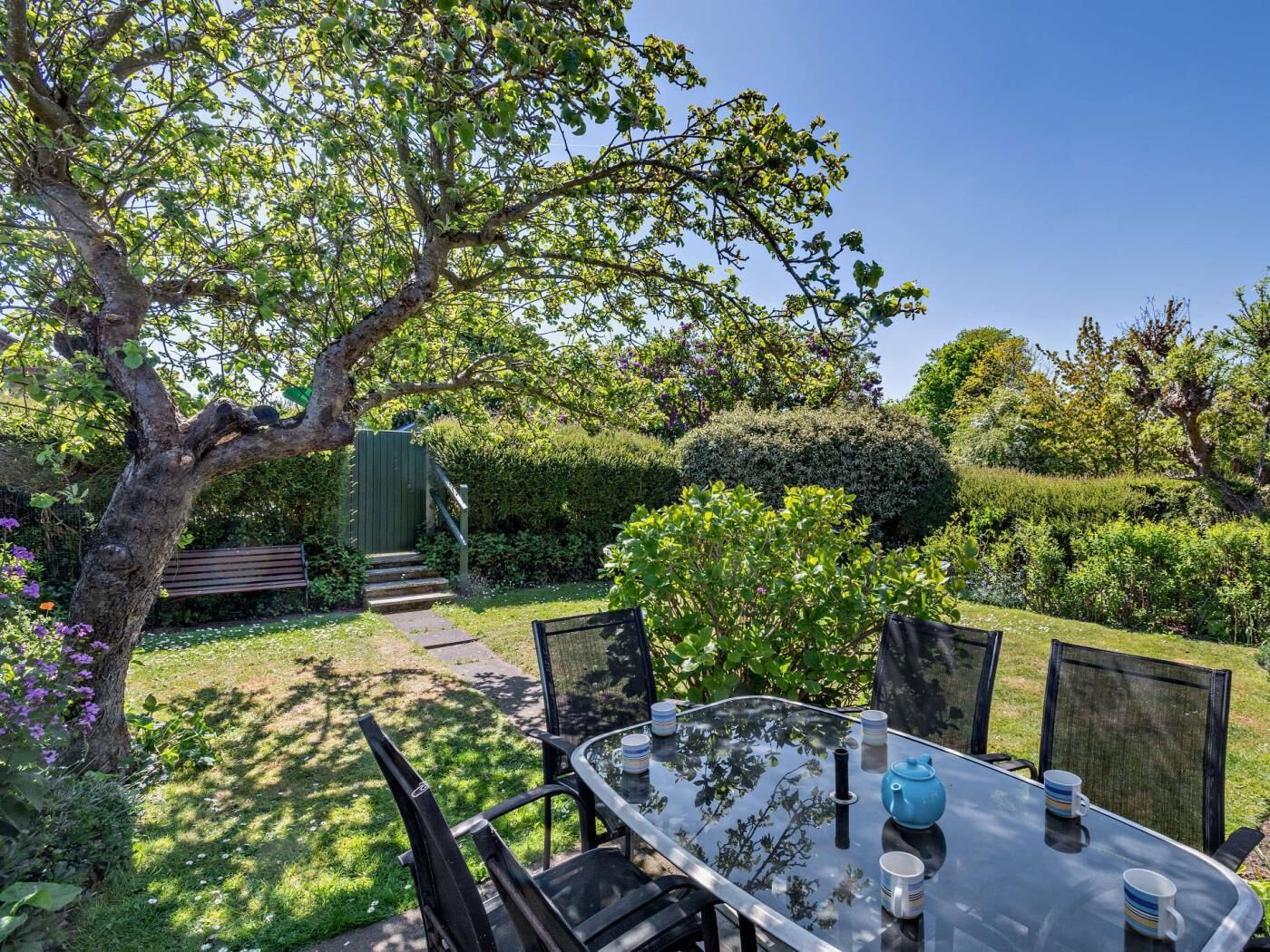 Ein Glastisch mit Stühlen, ein blauer Teekessel und Tassen in einem Garten.