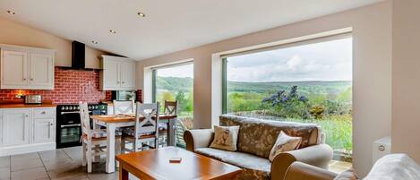 Cocina moderna con armarios blancos, salpicadero de ladrillo y comedor con mesa y sillas. Hay un sofá con vistas a un paisaje verde a través de amplias ventanas.