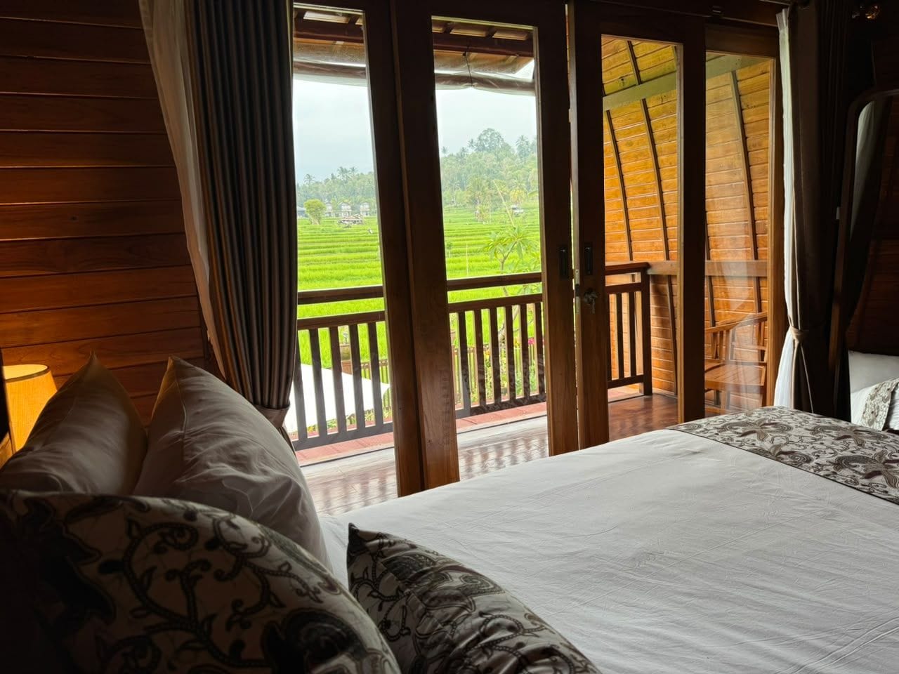 A room with a bed, a view of a green landscape through a sliding door, and a wooden balcony.