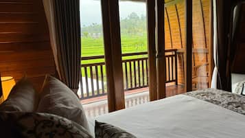 A room with a bed, a view of a green landscape through a sliding door, and a wooden balcony.
