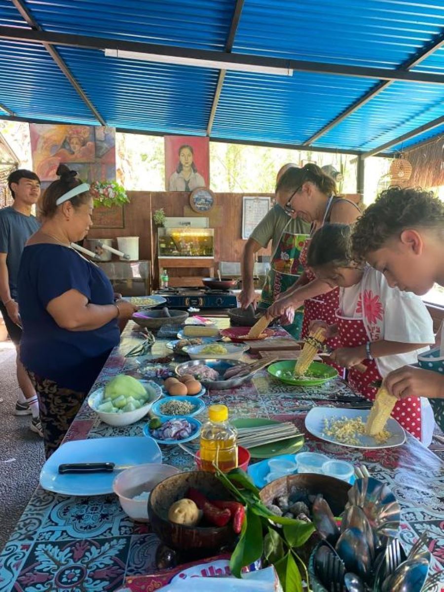 Un groupe de personnes prépare des aliments à une table installée à l’extérieur, en utilisant divers ingrédients et ustensiles de cuisine.