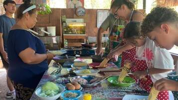 Un groupe de personnes prépare des aliments à une table installée à l’extérieur, en utilisant divers ingrédients et ustensiles de cuisine.