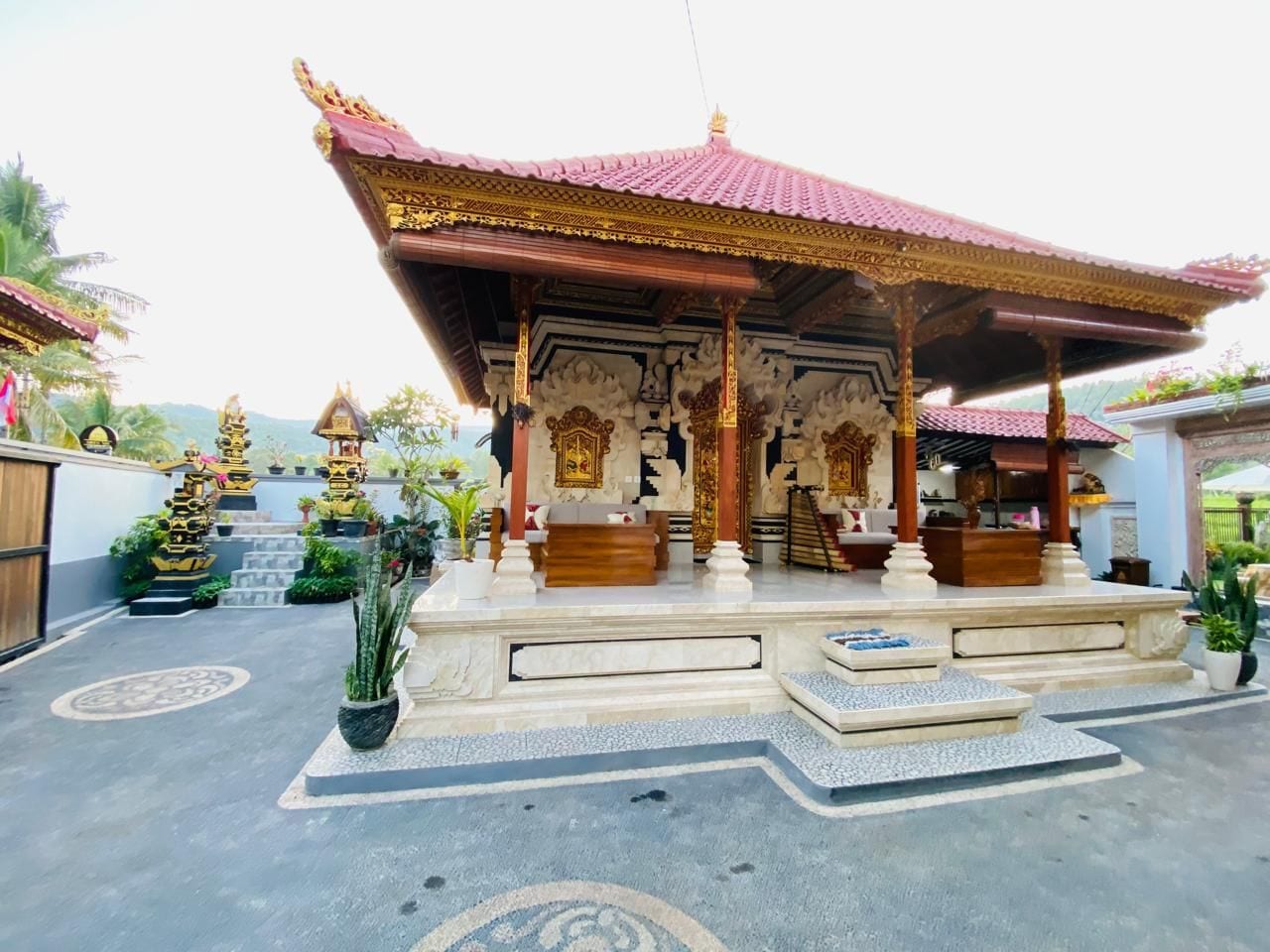 A traditional pavilion with ornate carvings and a tiled roof, surrounded by potted plants and a stone pathway.