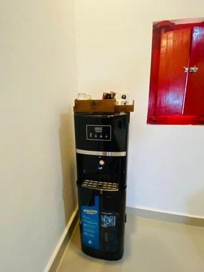 Eine GEALLEY Kaffeemaschine mit Wasserbehälter, platziert in einer Ecke eines Raumes mit einem roten Schrank und weißen Wänden.