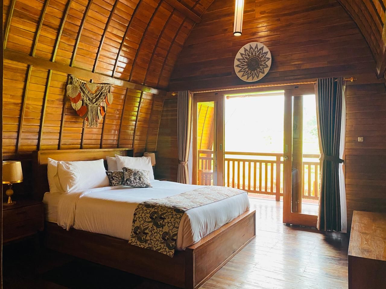 A wooden room with a bed, a nightstand, a wall hanging, and a view through a door.