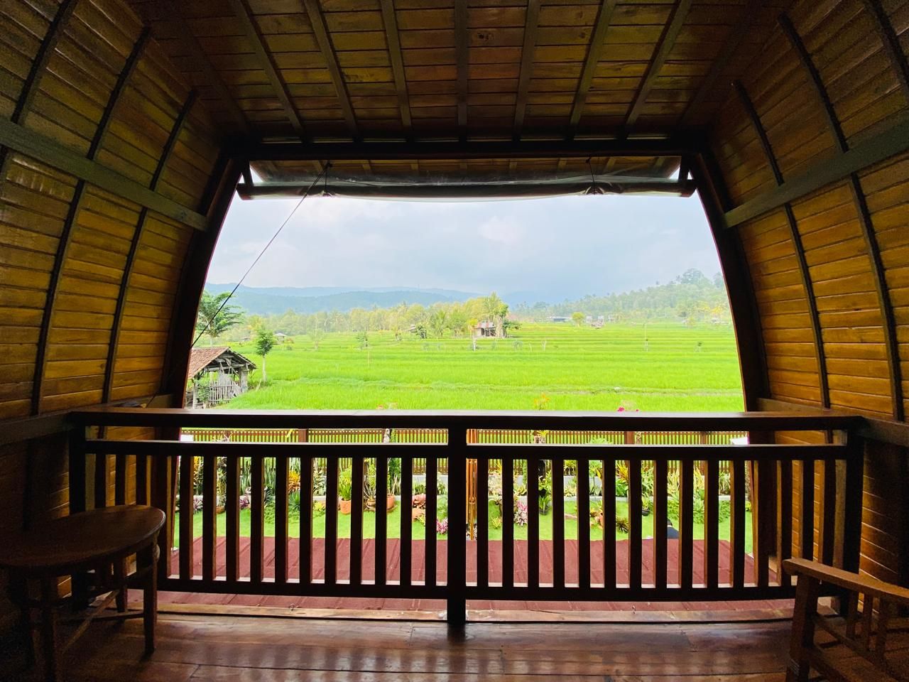 A wooden balcony with a view of a green field and a small house in the distance.