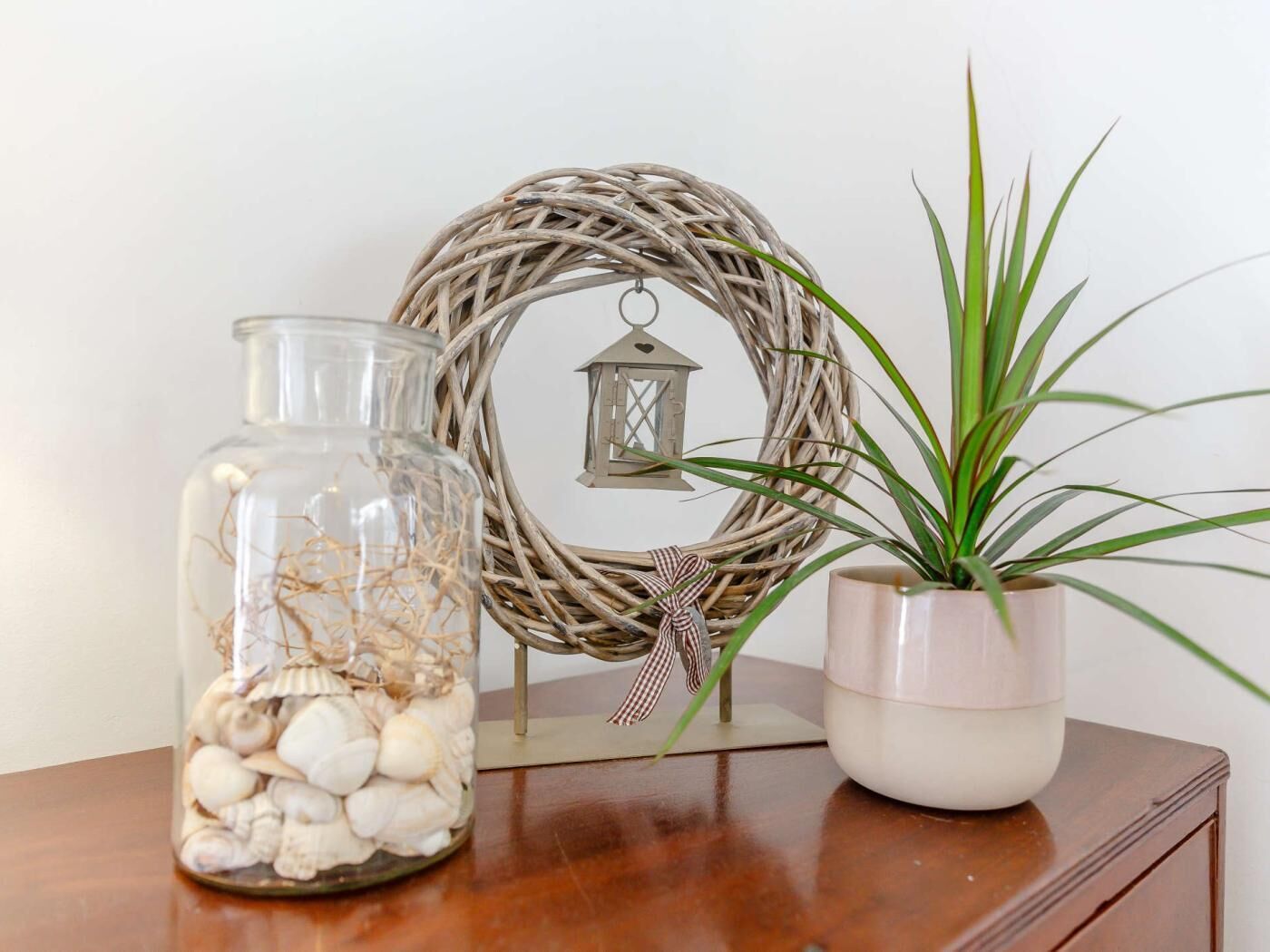 A glass jar filled with seashells, a potted plant, and a decorative wreath with a lantern on a wooden surface.