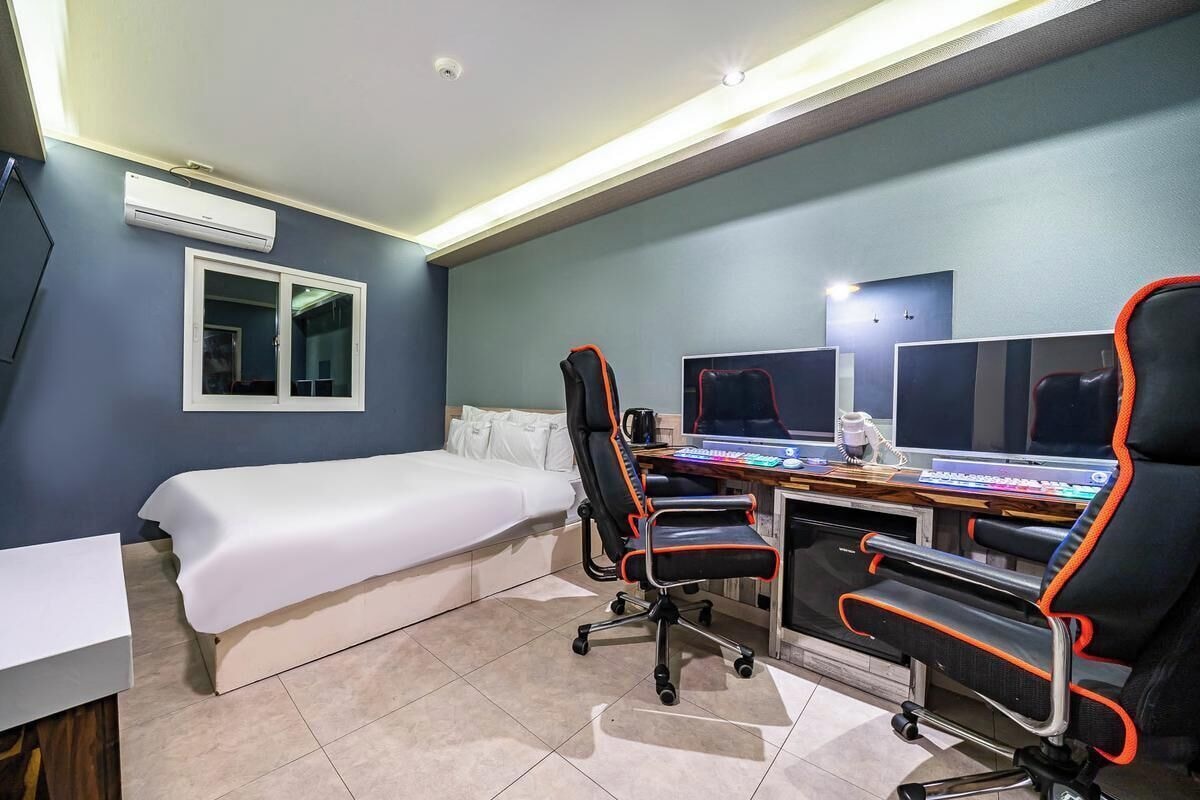 Camera d'albergo con un letto, una scrivania con due monitor, due sedie da ufficio, una TV e un condizionatore d'aria.