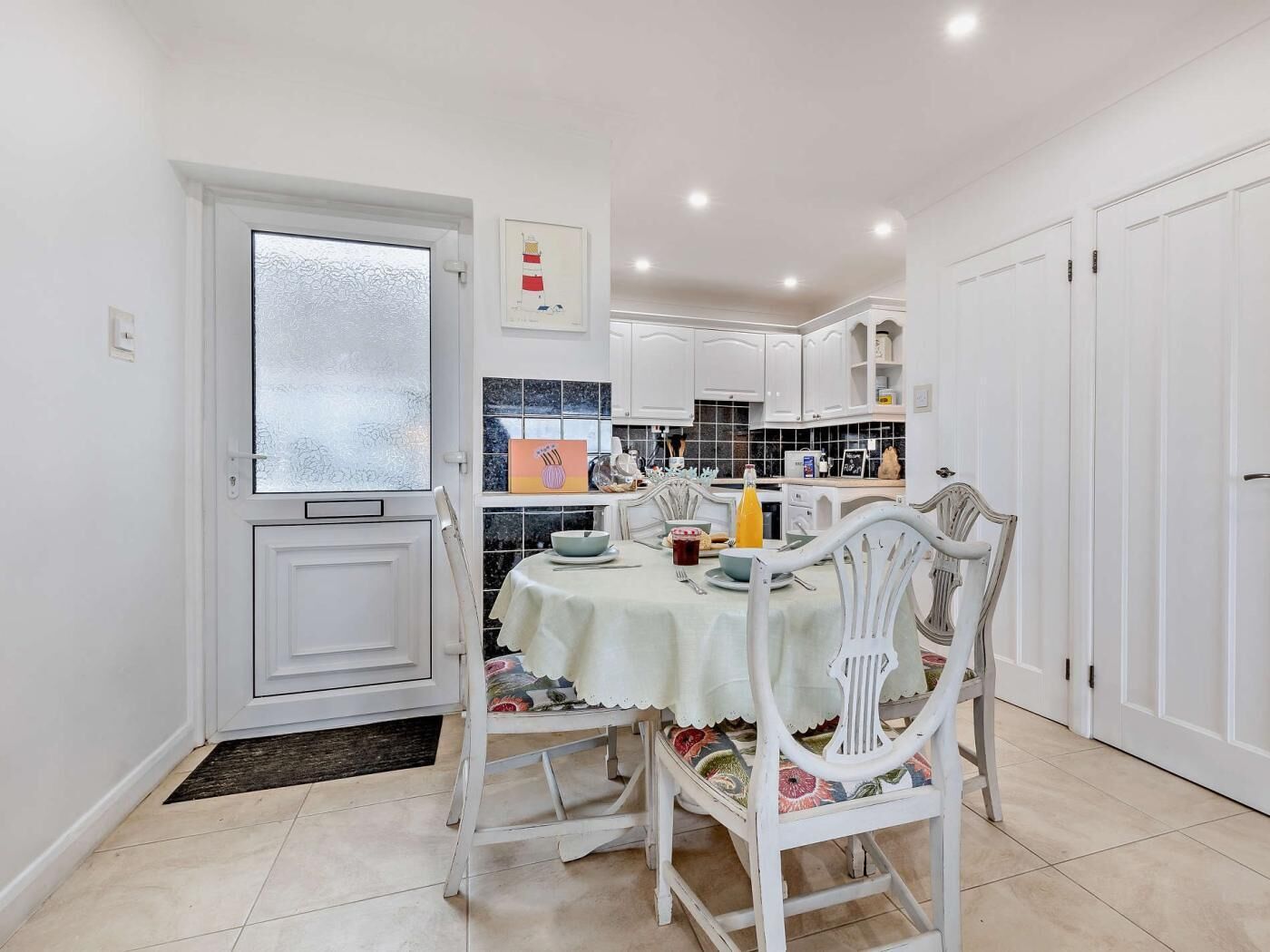 Un coin repas avec une table dressée pour deux personnes, des chaises blanches, une fenêtre à vitres dépolies et une cuisine en arrière-plan.