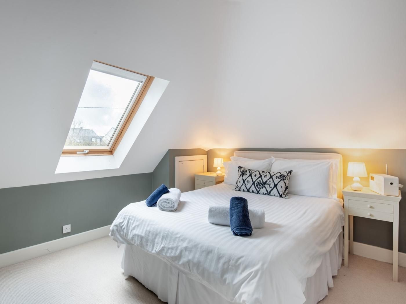 A bedroom with a skylight, a bed with white bedding, two bedside tables with lamps, and a patterned pillow.