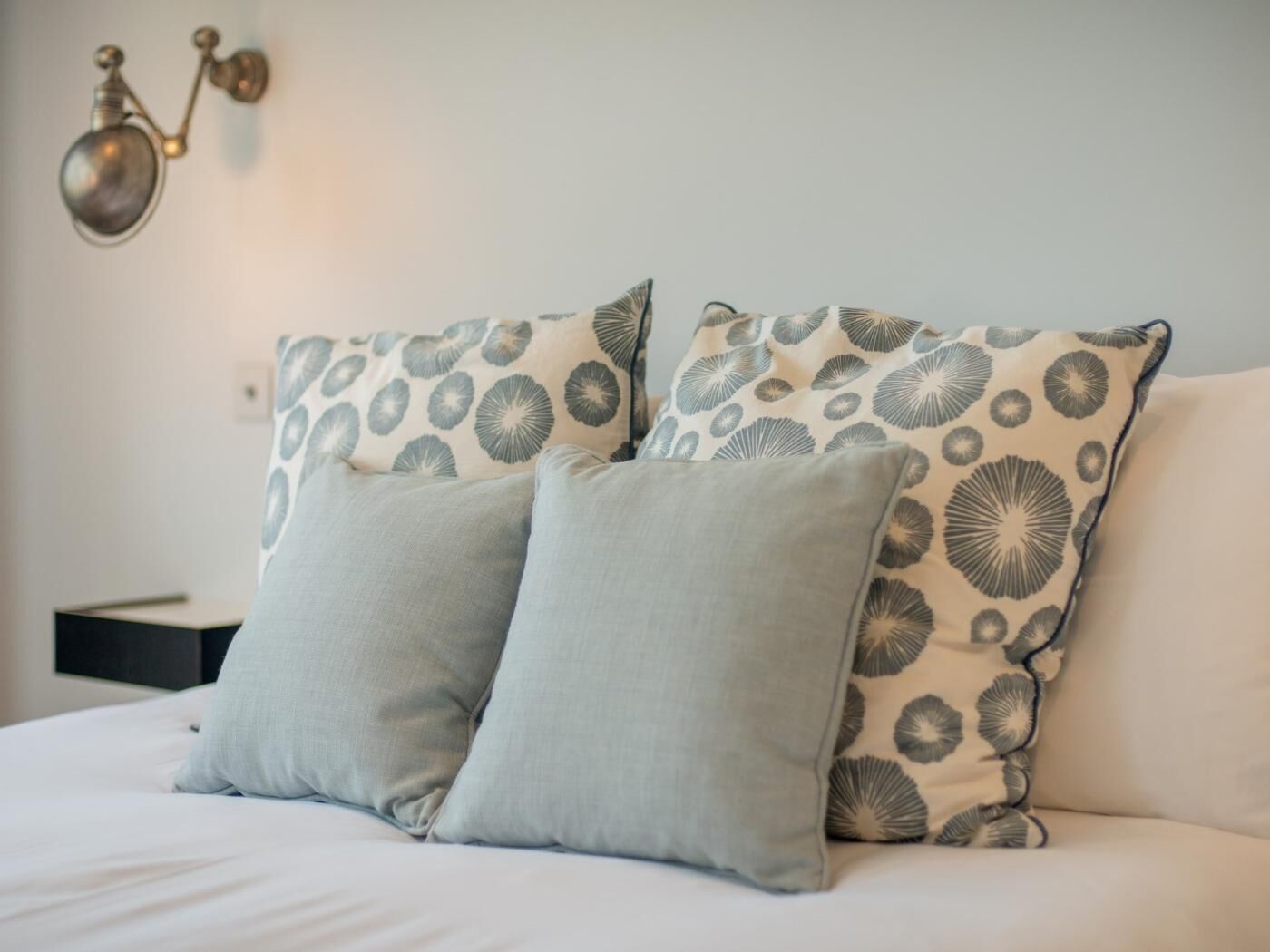 Una cama con dos almohadas, una con estampado floral y otra lisa, y una lámpara de pared.