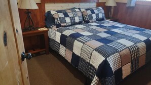 A bedroom with a wooden headboard, a quilted bedspread, two bedside lamps, and a wooden nightstand. - Alpha Wolf Cabin at the Wolf Haven Cabins (Lovell)