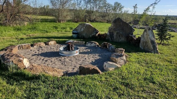 A circular stone fire pit with a metal bowl in the center, surrounded by rocks and grass. - Alpha Wolf Cabin at the Wolf Haven Cabins (Lovell)