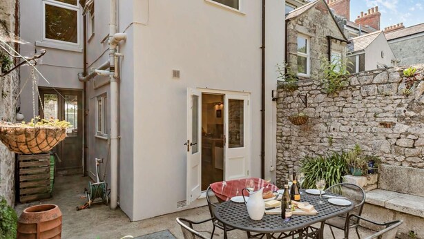 A courtyard with a dining table, chairs, a wine bottle, and a basket of fruit. - 3 Bed in Portland (oc-dc031) (Portland)