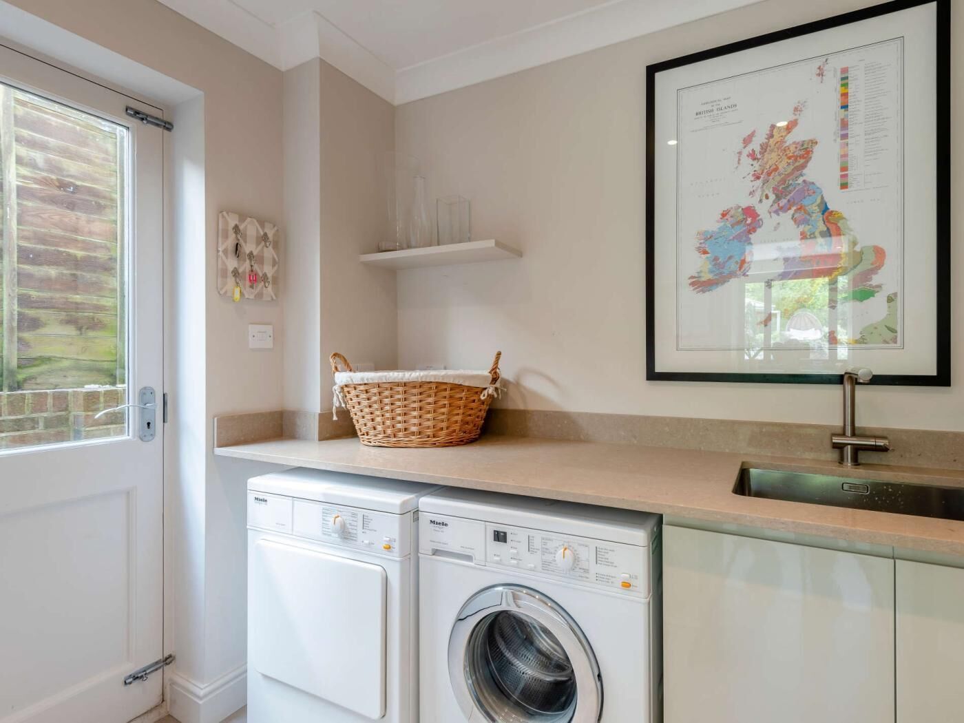 A laundry area with two Miele washing machines, a wicker basket, a framed map, and a window with a view of greenery.