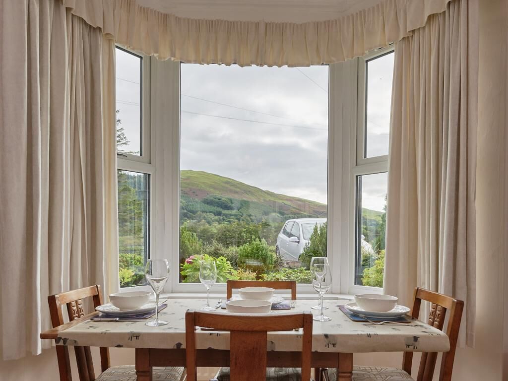 A dining table with chairs and a view of a car and hills through large windows.