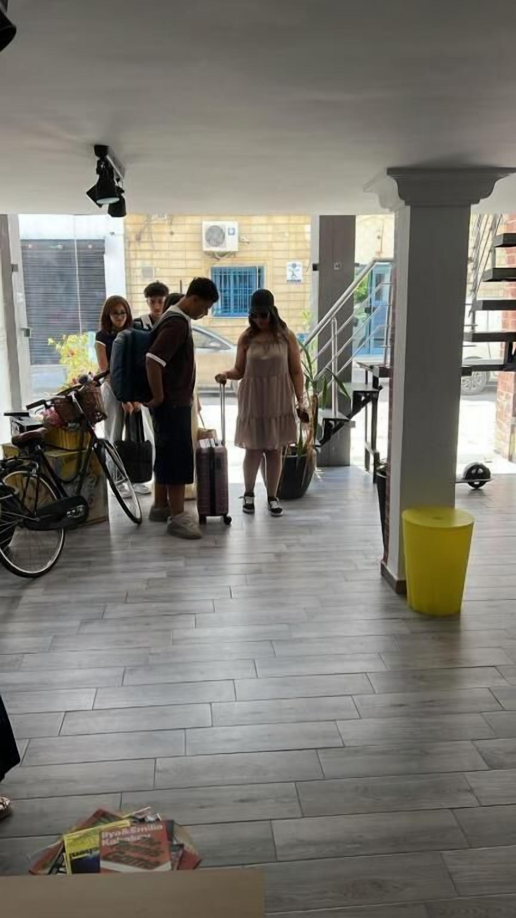 Un grupo de personas cerca de una escalera, algunas con equipaje, en un edificio con piso de baldosas y un bote de basura amarillo.