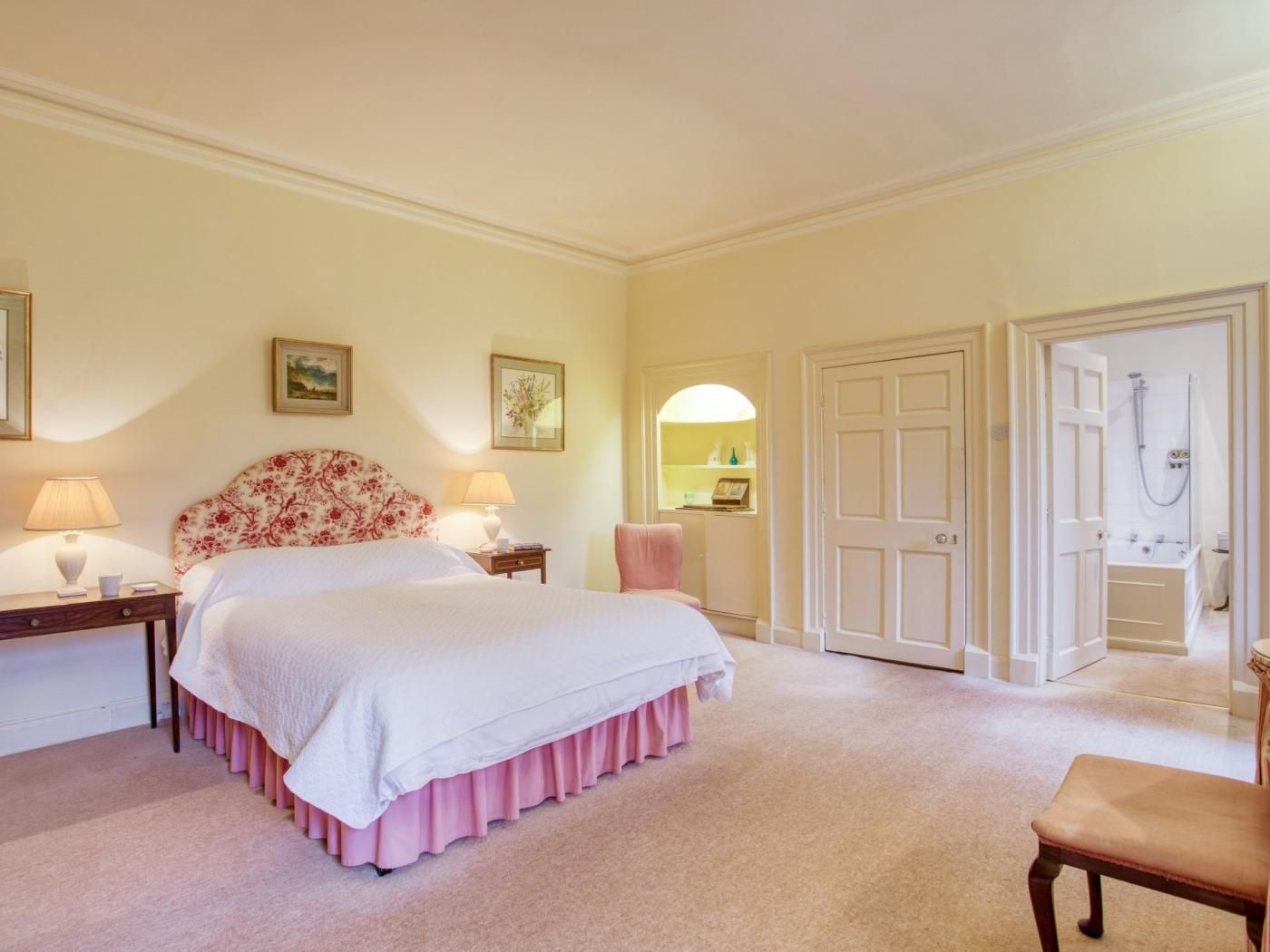 A bedroom with a bed, two bedside tables with lamps, a chair, a door leading to a bathroom, and a wall with two framed pictures.