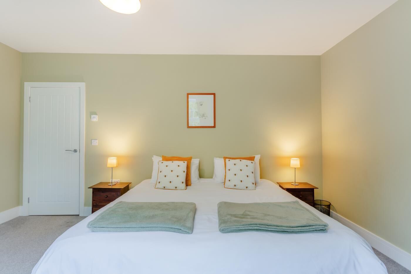 A hotel room with a bed, two bedside tables, a nightstand, a door, and a framed picture on the wall.