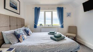 Um quarto com cama, janela com cortinas, uma cadeira e televisão de tela plana.