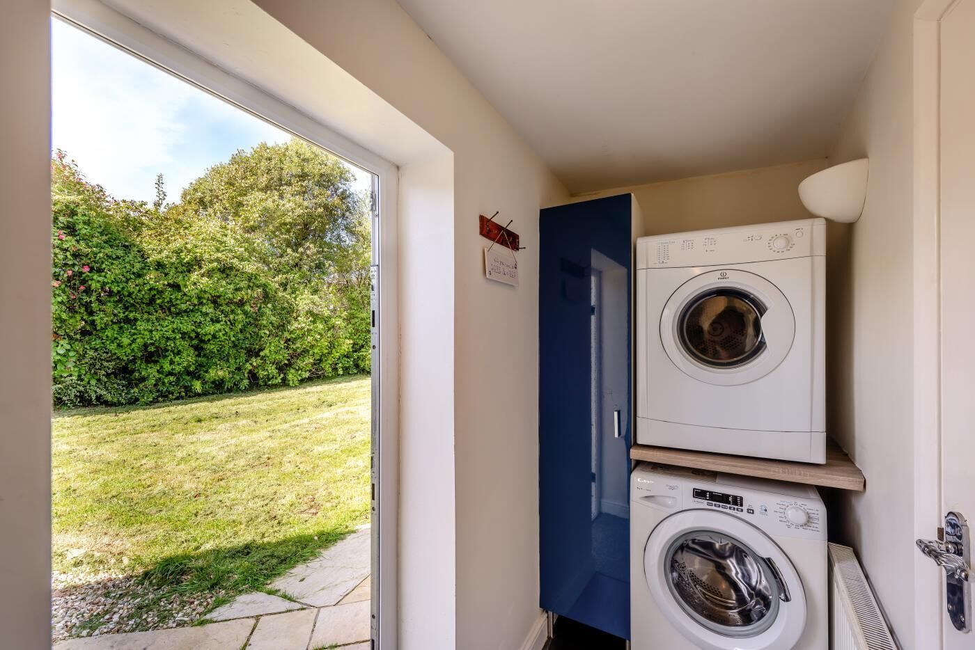 A laundry room with two stacked washing machines, a blue door, and a view of a grassy yard through a glass door.