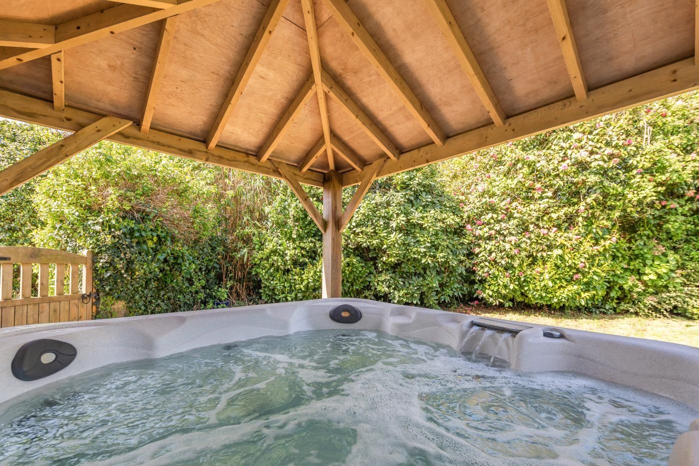 A hot tub under a wooden pergola with a garden backdrop.