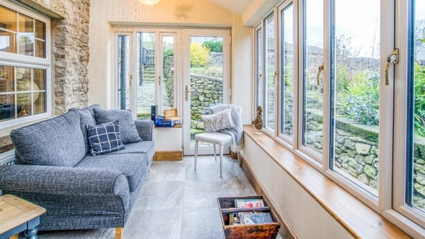 A cozy room with a stone wall, a grey sofa, a white chair, and a wooden table. - 3 Bed in Buxton (oc-77739) (Hartington)