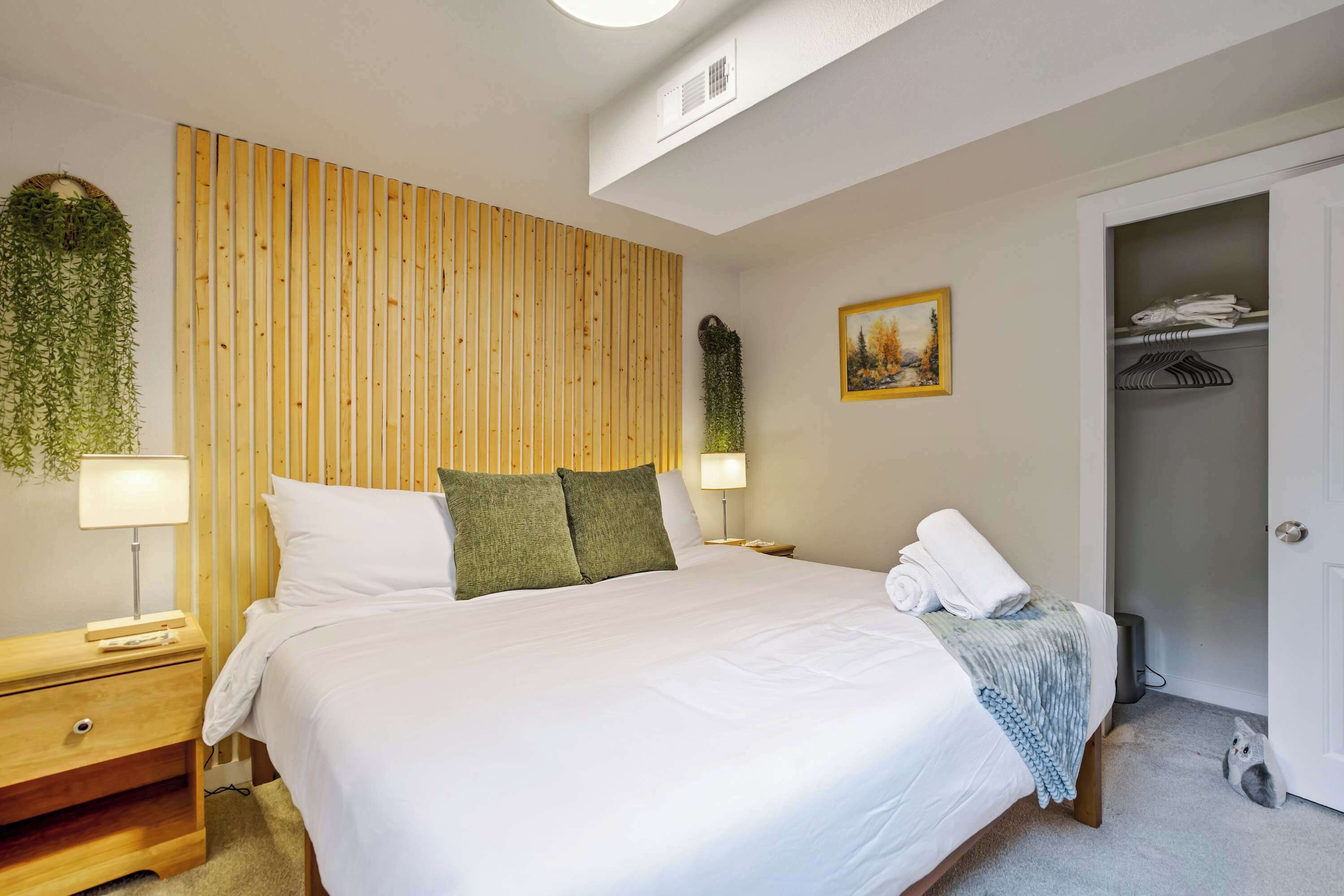 Un dormitorio ordenado con cama, mesitas de noche, una planta en la pared, un cuadro y un armario.