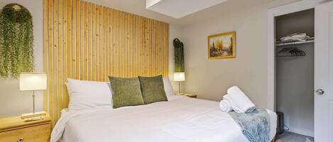 Un dormitorio ordenado con cama, mesitas de noche, una planta en la pared, un cuadro y un armario.
