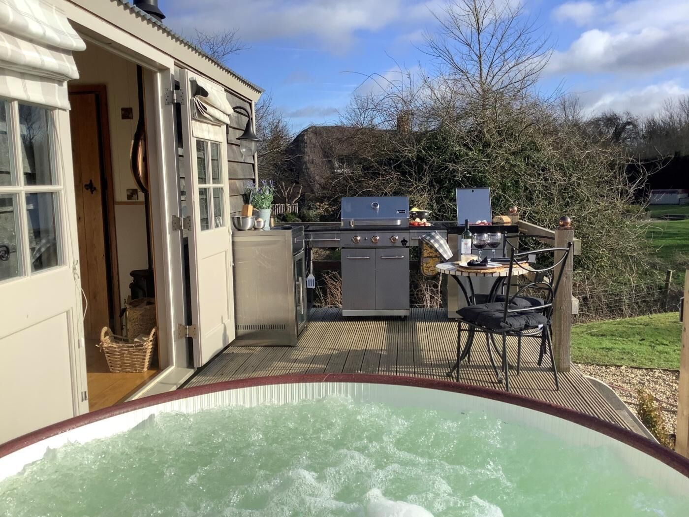 A hot tub with a wooden deck, a barbecue grill, and a dining table with chairs.