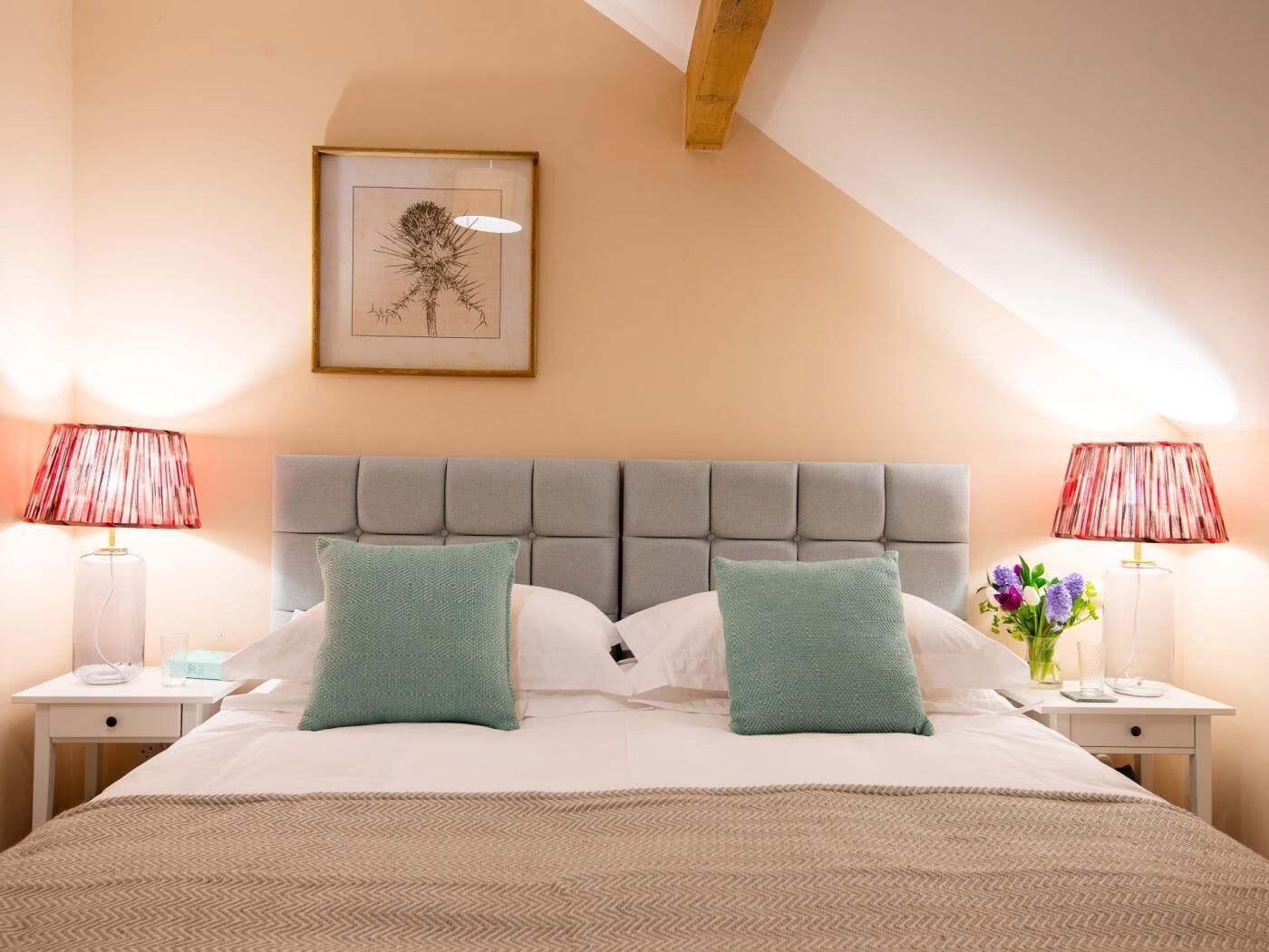 Een net opgemaakt bed met twee kussens, een nachtkastje met een lamp, en een lijst met kunst aan de muur.