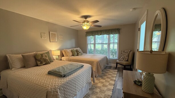 A bedroom with two beds, a ceiling fan, a window with blinds, a chair, a lamp, and a mirror. - Fairway Vista - Newly Renovated with Beautiful Golf Course views, 1bed-1 bath (Pawleys Island)