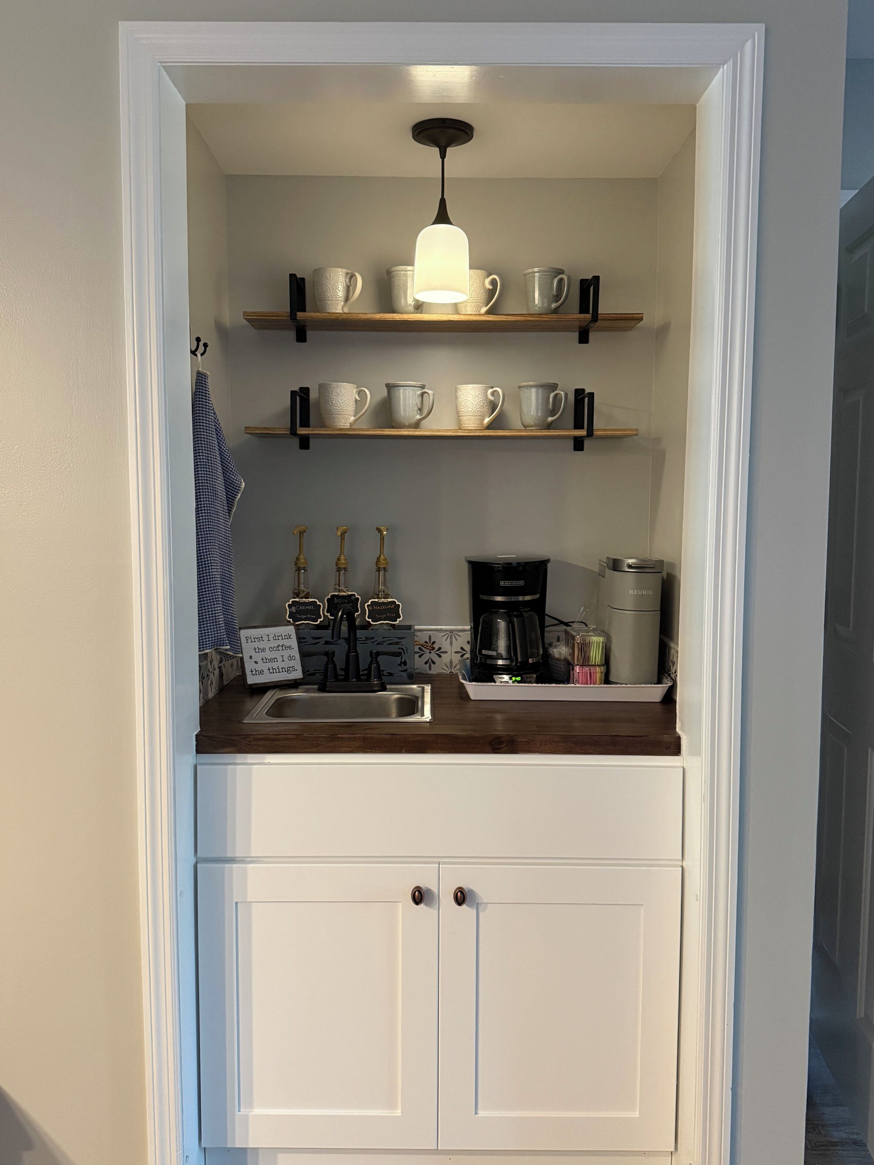 A compact kitchen with a built-in sink, a coffee maker, and shelves holding cups and mugs.
