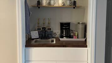 A compact kitchen with a built-in sink, a coffee maker, and shelves holding cups and mugs.