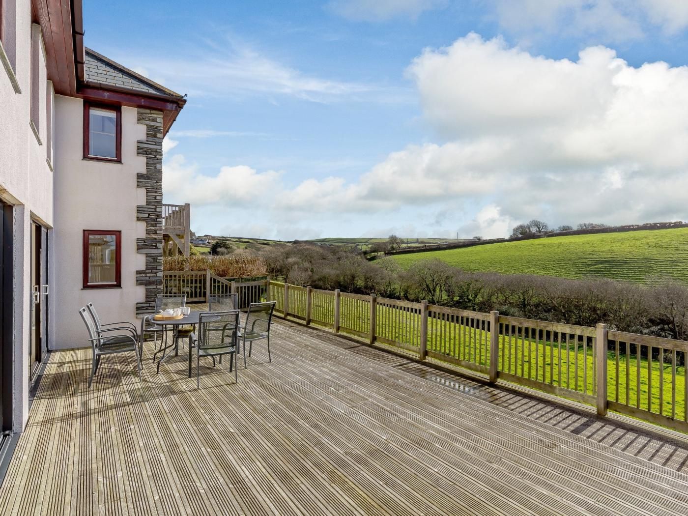 A wooden deck with outdoor furniture, a stone wall, and a view of green fields.