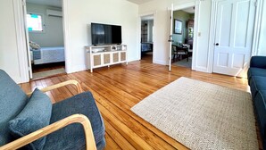 A spacious living room with a wooden floor, a television on a white cabinet, a sofa, and a patterned rug.