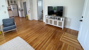 A living room with a TV, wooden flooring, a sofa, and a kitchen in the background.