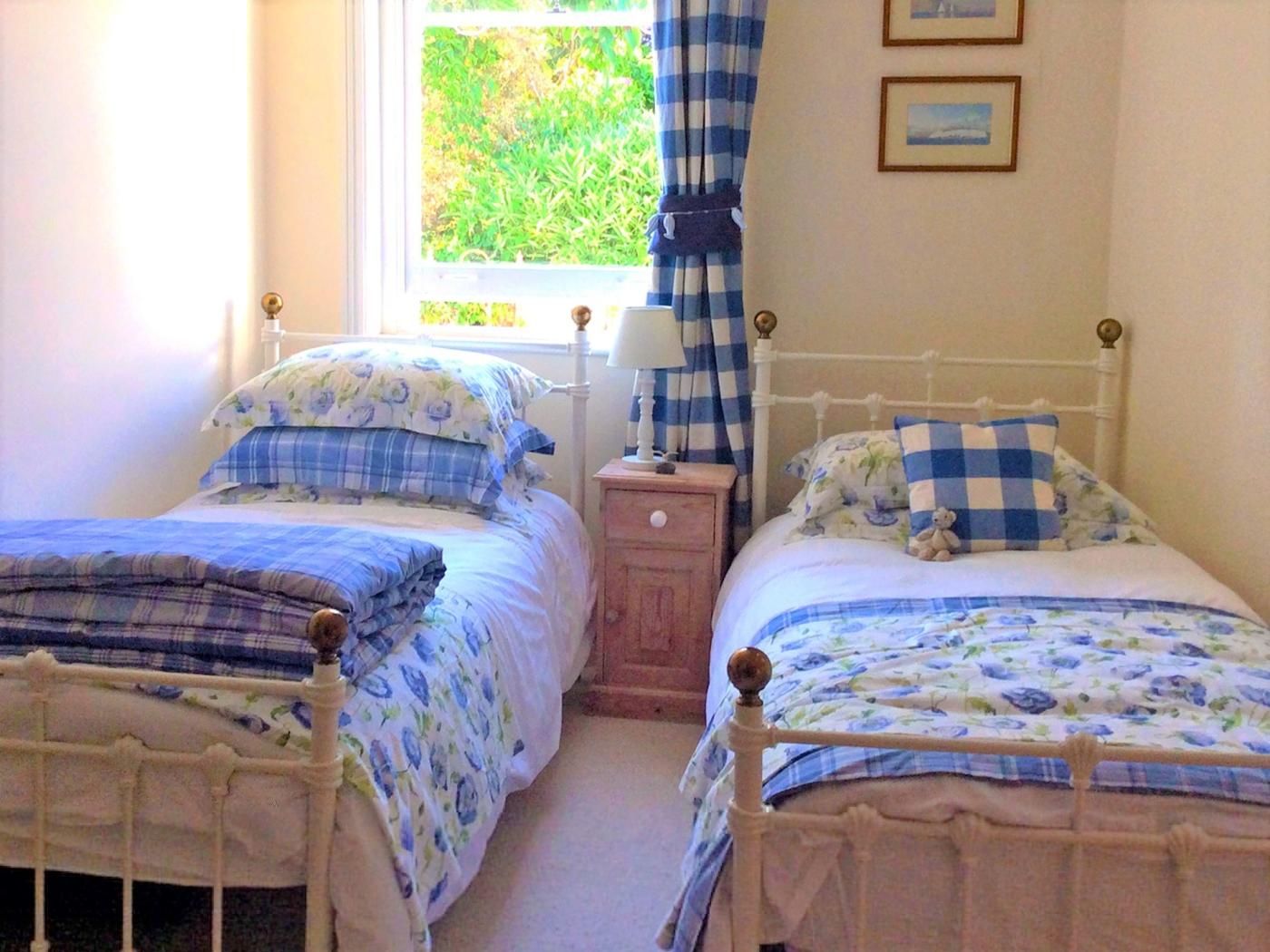 Dos camas individuales con ropa de cama floral, una mesita de noche con lámpara, una ventana con cortinas e imágenes enmarcadas en la pared.