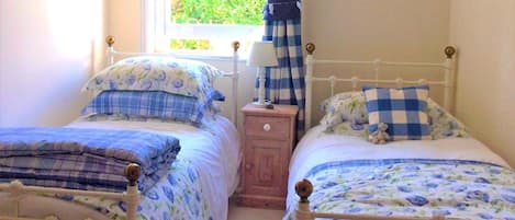 Dos camas individuales con ropa de cama floral, una mesita de noche con lámpara, una ventana con cortinas e imágenes enmarcadas en la pared.