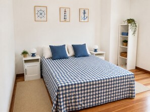 A neatly arranged bedroom with a checkered bedspread, bedside tables with lamps, a nightstand with a plant, and a white shelving unit with a plant.