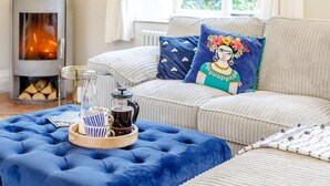 Een gezellige woonkamer met een bank, een blauwe poef, een salontafel met dienblad en een open haard.