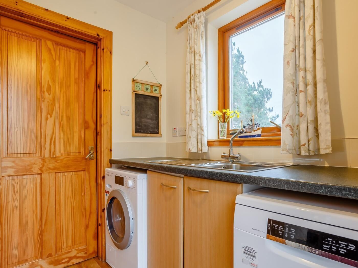 Cocina compacta con armarios de madera, lavadora, lavavajillas y una ventana con cortinas.