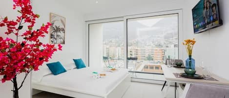 A modern bedroom with a bed, a dining area, and a large window with a city view.