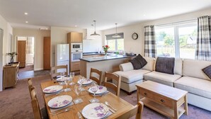 Eine moderne Küche mit eingebauten Geräten, ein Essbereich mit einem Holztisch, gedeckt für vier Personen, und ein Wohnbereich mit einer Couch und einem Fenster mit Blick auf einen Garten.